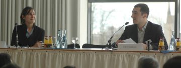 Mann und Frau nebeneinander an einem langen Tisch vor Silhouette des Publikums