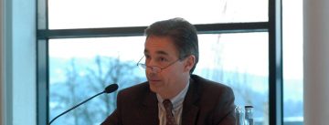 Mann im grauen Sakko schaut über den Rand seiner Brille hinweg, er hält einen Papierstapel