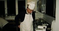 Koch steht vor einer Küchenmaschine, er bindet sich eine weiße Schürze an, Quelle: DTF