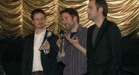 drei Männer mit Mikrofone in den Händen vor goldenem Vorhang, Quelle: DTF