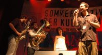 Band auf rot beleuchteter Bühne vor Banner des Sommerfestivals, Quelle: DTF