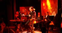 Sänger sitzt auf orange beleuchteter Bühne zwischen verschiedenen Musikern, Quelle: DTF
