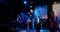 Sänger sitzt auf blau beleuchteter Bühne zwischen verschiedenen Musikern, Quelle: DTF