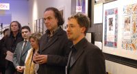 Menschen stehen vor Wand mit Fotos, Kerim Arpad spricht mit einer Moderationskarte in den Händen, Quelle: DTF