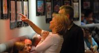 Mann und Frau vor Wand mit Fotos, die Frau zeigt mit dem rechten Zeigefinger, Quelle: DTF
