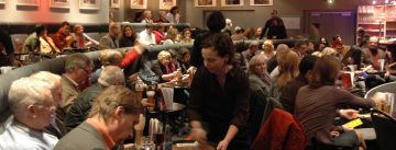Menschen auf bequeme Bänke mit Getränke, Kellner*innen gehen von Tisch zu Tisch