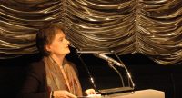 Frau mit buntem Schal spricht am Rednerpult vor goldenem Vorhang, Quelle: DTF