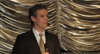 junger Mann mit grüner Krawatte spricht am Rednerpult vor goldenem Vorhang, Quelle: DTF