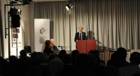 Diskussionteilnehmer*innen, neben ihnen spricht ein Mann am Rednerpult vor Silhouette des Publikums, Quelle: DTF