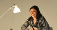 Frau lächelnd an einem Holztisch mit darüber hängender Schreibtischlampe, Quelle: DTF