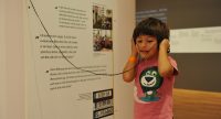 Junge mit Kopfhörer um den Kopf vor einer Ausstellungswand, Quelle: DTF
