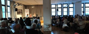 zwei Männer und eine Frau sitzend an Holztischen vor Banner des DTF vor Publikum