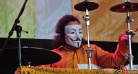 Mann in orangenem Hemd und schwarz-weißer Maske spielt Schlagzeug, Quelle: DTF