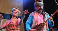 Mann mit traditionellem Kopfschmuck spielt Saiteninstrument, hinter ihm tanzt eine Frau, Quelle: DTF