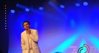 Mann im weißen Anzug steht auf blau beleuchteter Bühne vor Silhouette des Publikums, Quelle: DTF
