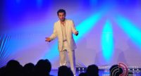 Mann im weißen Anzug steht auf blau beleuchteter Bühne vor Silhouette des Publikums, Quelle: DTF