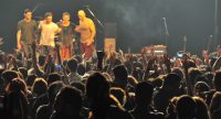 Menschen mit erhobenen Händen vor Bühne mit vier Musikern in freundschaftlicher Umarmung, Quelle: DTF