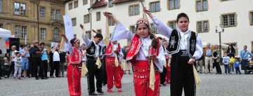 Mädchen und Jungs in traditioneller Kleidung stehen auf dem Platz mit schräg erhobener rechter Hand und linker Hand an der Hüfte