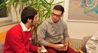 zwei junge Männer sitzen sprechend nebeneinander mit Kaffeetassen in den Händen, Quelle: DTF