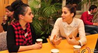 zwei junge Frauen sprechen am Holztisch nebeneinander mit Kaffeetassen vor sich, Quelle: DTF