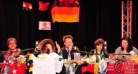 drei Frauen und zwei Männer stehend und sitzend an einem dicht bedeckten roten Tisch mit Länderflaggen vor SIlhouette des Publikums, Quelle: DTF