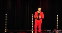Mann im roten Anzug spricht gestikulierend auf der Bühne vor Silhouette des Publikums, Quelle: DTF