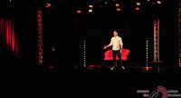 Mann im weißen Hemd gestikulierend auf der Bühne vor einem roten Sofa vor Silhouette des Publikums, Quelle: DTF