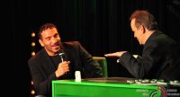 zwei Männer sitzend an einem grünen Schreibtisch unterhalten sich vor Silhouette des Publikums, Quelle: DTF