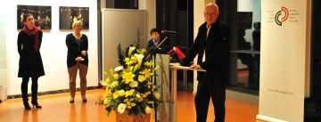 Mann an Rednerpult vor Banner des DTF in einem Ausstellungsraum, vor ihm steht eine Vase mit gelben Blumen