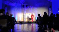 Mann im roten Anzug auf blau beleuchteter Bühne vor Silhouette des Publikums, Quelle: DTF