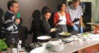 zwei Frauen und ein Mann stehen an einem Tisch mit Tellern drauf, sie kochen, während links ein Journalist mit grünem Mikrofon steht, Quelle: DTF