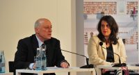 Mann im schwarzen Sakko an Holztisch sitzend neben Frau im weißen Sakko, Quelle: DTF