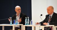 zwei Männer in schwarzen Anzügen sitzend an Holztischen sprechen miteinander, Quelle: DTF