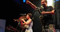 Saxophonist zeigt mit rechter Hand ins Publikum, Trompeterin und Akkordeonspieler im Hintergrund, Quelle: DTF