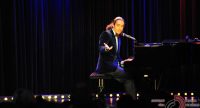Mann mit langen Haaren im dunkelvioletten Sakko sitzt an einem Klavierflügel und zeigt ins Publikum, Quelle: DTF