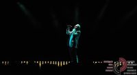 Mann in weißer Kleidung steht im dunklen Saal türkis beleuchtet auf der Bühne und singt, Quelle: DTF