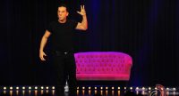 Mann im schwarzen Shirt steht vor einem pinken SOfa auf der Bühne vor SIlhouette des Publikums, Quelle: DTF