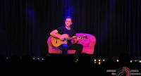 Mann im schwarzen Shirt sitzt auf einem pinken SOfa auf der Bühne vor SIlhouette des Publikums und spielt Gitarre, Quelle: DTF