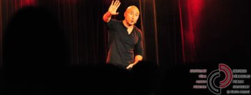 Mann im schwarzen Shirt steht mit erhobener rechter Hand auf der Bühne