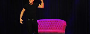 Mann im schwarzen Shirt steht vor einem pinken SOfa auf der Bühne vor SIlhouette des Publikums