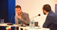zwei Männer sitzend an Holztischen mit Banner des DTF im Hintergrund, Quelle: DTF