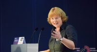 blonde Frau in dunkelblauem Hemd spricht gestikulierend am Rednerpult mit Blick Richtung Publikum, Quelle: DTF