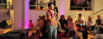 Mann im pinken Shirt steht mitten im Publikum mit Mikrofon in der Hand