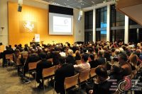 Quelle: DTF volle Sitzreihen im hölzernen Saal schauen sprechender Frau am Rednerpult zu, Quelle: DTF