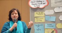 Frau vor Pinnwand mit beschrifteten Karteikarten spricht gestikulierend, Quelle: DTF
