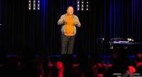 Mann im orangen Hemd gestikuliert lachend Richtung SIlhouette des Publikums, Quelle: DTF