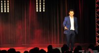 Mann im blauen Anzug auf rot beleuchteter Bühne vor Silhouette des Publikums, Quelle: DTF