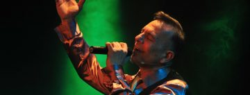 Mann im glänzenden Hemd hebt die rechte Hand nach oben während er auf grün beleuchteter Bühne singt