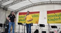 Mann in gelbem T-SHirt und Frau in schwarzem T-SHirt auf weißer Bühne vor Bannern des Interkulturellen Kinderfestes, der Mann spricht ins Mikrofon, Quelle: DTF