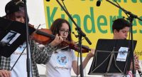 zwei Jungs und ein Mädchen in weißen Shirts spielen Musik vor Banner des Kinderfestes, Quelle: DTF